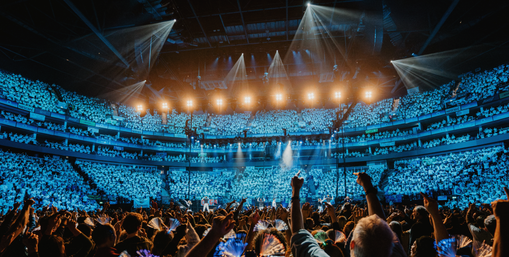 A photo of the O2 stage with audience with their arms raised.